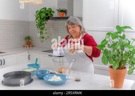 Femme chef dans un tablier blanc battant des oeufs dans un bol avec un mélangeur électrique pour faire un gâteau de fraise Banque D'Images