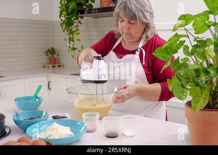 Femme chef dans un tablier blanc battant des oeufs dans un bol avec un mélangeur électrique pour faire un gâteau de fraise Banque D'Images