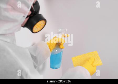 Femme en costume de protection nettoyant moule avec pulvérisateur et chiffon sur le mur à l'intérieur, gros plan Banque D'Images