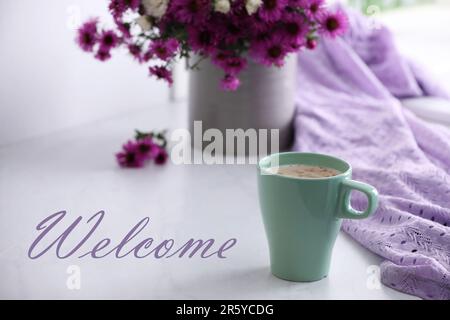 Carte de bienvenue. Tasse de café aromatique, belles fleurs et tissu violet sur table blanche Banque D'Images