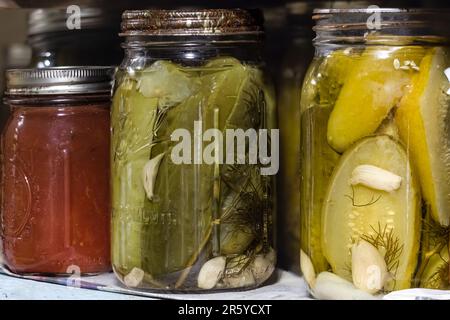 Deux bocaux de mise en conserve remplis de cornichons verts et de gousses d'ail, et un pot de mise en conserve rempli de sauce tomate rouge placé sur une étagère dans un sous-sol Banque D'Images