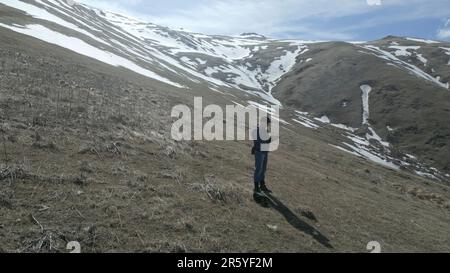 Un jeune homme se tient dans les hautes montagnes, couvert d'herbe verte et de griffes enneigées, un sommet de falaise enneigée. Un jour ensoleillé contre un ciel bleu avec du whi Banque D'Images