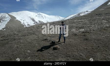 Un jeune homme se tient dans les hautes montagnes, couvert d'herbe verte et de griffes enneigées, un sommet de falaise enneigée. Un jour ensoleillé contre un ciel bleu avec du whi Banque D'Images