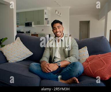 Joyeux jeune homme biracial dans des vêtements décontractés assis sur un canapé dans le salon à la maison Banque D'Images