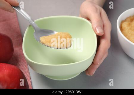 Une alimentation saine pour bébé. Femme mettant une délicieuse purée de pommes dans un bol à table gris clair, gros plan Banque D'Images