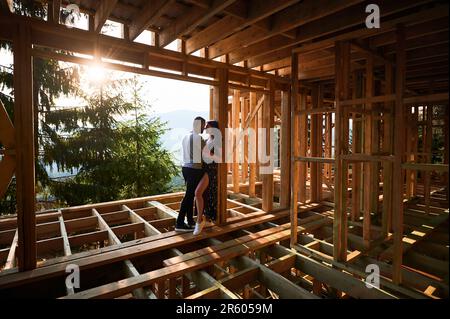 Homme et femme examinant leur futur logement en bois de cadre niché dans les montagnes près de la forêt. Jeune couple sur le chantier en début de matinée. Concept de construction écologique contemporaine. Banque D'Images