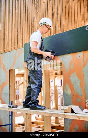 Carpenter construisant une maison en bois encadrée. Homme ouvrier en lunettes habillage façade de maison avec panneaux de particules de ciment, les fixant avec des tournevis. Concept de construction écologique moderne. Banque D'Images