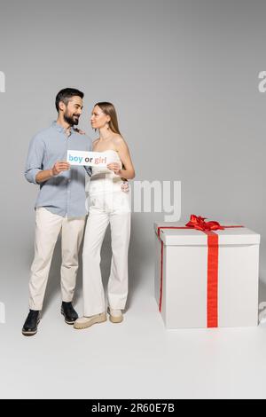 Souriant et élégant s'attendant à des parents tenant la carte avec le jeune garçon ou fille lettrage et embrassant près de grande boîte cadeau pendant le sexe révéler surprise partie sur gre Banque D'Images