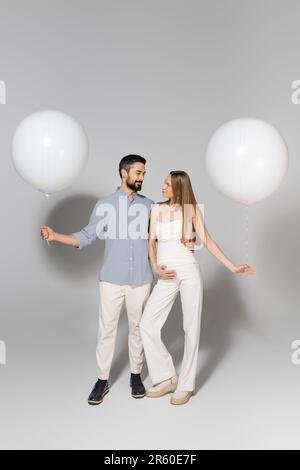 Une longueur complète de parents à la mode s'embrassant et regardant les uns les autres tout en tenant des ballons de fête pendant le sexe révèle la fête surprise sur g Banque D'Images