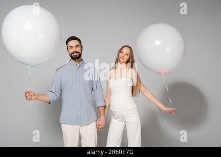 Souriant et élégant, s'attendant à ce que les parents tiennent les mains et les ballons blancs festifs tout en regardant l'appareil photo pendant le sexe révèlent la fête surprise sur bac gris Banque D'Images