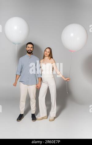 Pleine longueur de positif et élégant s'attendant à des parents regardant la caméra tout en tenant les mains et ballons pendant le sexe révéler la fête surprise sur le b gris Banque D'Images
