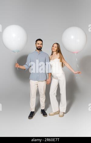 Pleine longueur de parents à la mode et heureux s'attendant à tenir des ballons blancs et regardant la caméra pendant le sexe révéler la fête surprise sur fond gris Banque D'Images