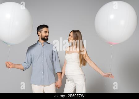 Souriant et tendance, s'attendant à ce que les parents tiennent les mains et les ballons blancs de fête tout en regardant les uns les autres pendant la fête et le sexe révèle des surprenis Banque D'Images