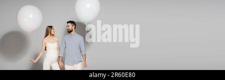 Femme enceinte à la mode tenant la main d'un mari souriant et ballon de fête pendant le sexe révèle une fête surprise sur fond gris avec espace pour les copies, Banque D'Images