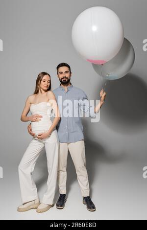 Pleine longueur d'homme positif et élégant tenant des ballons de fête et embrassant femme enceinte pendant la fête de douche de bébé sur fond gris, s'attendant à la pare Banque D'Images