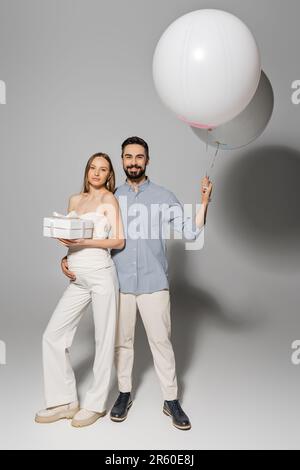 Pleine longueur d'homme positif tenant des ballons festifs et embrassant la femme branchée et enceinte avec boîte cadeau tout en se tenant ensemble pendant la douche de bébé par Banque D'Images