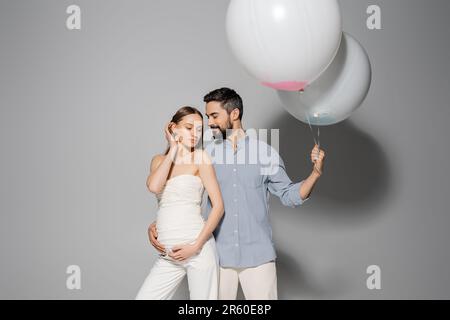 Homme souriant et barbu embrassant une femme enceinte élégante et tenant des ballons festifs pendant la fête des femmes et la fête sur fond gris, en attendant p Banque D'Images