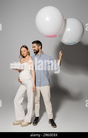 Homme gai embrassant une femme enceinte élégante et tenant une boîte cadeau et des ballons festifs pendant la fête et le genre révèlent une fête surprise sur fond gris Banque D'Images