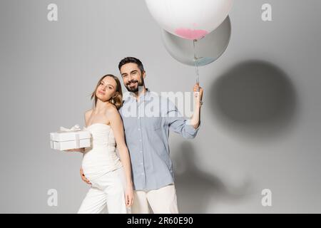 Homme positif et élégant tenant des ballons de fête et embrassant femme enceinte avec boîte cadeau pendant la fête et le sexe révèle la fête surprise sur gris Banque D'Images