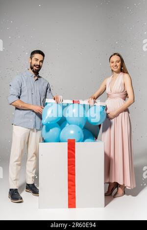 Toute la longueur de couple tendance regardant l'appareil photo tout en ouvrant la boîte cadeau avec des ballons bleus et debout sous des confettis pendant la douche de bébé sur le dos gris Banque D'Images