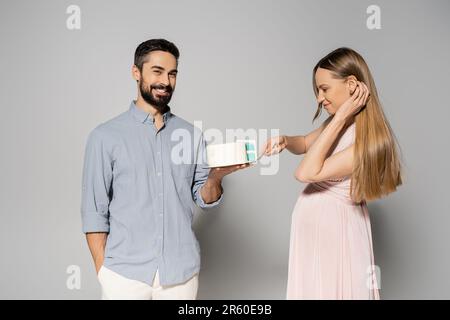 Homme positif et barbu tenant le gâteau près de l'élégante femme enceinte en robe rose pendant la fête de genre sur fond gris, s'attendant parents concept, gend Banque D'Images