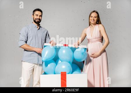 Femme enceinte positive dans une élégante robe ouverture bog boîte cadeau avec ballons bleus tout en se tenant près de mari sous confetti pendant la douche de bébé sur gris Banque D'Images