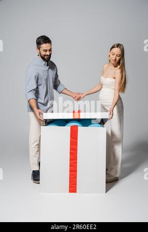 Un homme positif tenant la main de la femme enceinte tout en ouvrant la boîte cadeau avec des ballons bleus pendant le sexe révèle la fête surprise sur fond gris, s'attendant Banque D'Images