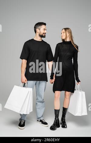 Homme barbu souriant tenant des sacs de shopping et regardant la femme enceinte à la mode en robe et debout sur fond gris, de nouveaux débuts et parent Banque D'Images