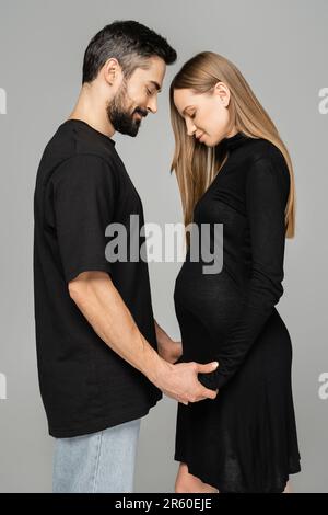 Vue latérale de l'homme barbu en jeans et t-shirt tenant les mains d'une femme à poil long, élégante et enceinte tout en se tenant isolé sur des débuts gris Banque D'Images