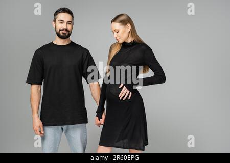 Homme barbu souriant en t-shirt et jeans tenant main de femme élégante et enceinte en robe noire et debout isolé sur gris, concept d'attente Banque D'Images