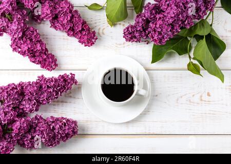 Tasse de café noir frais et branche de lilas violets fleuris avec feuilles vertes sur fond de bois blanc. Pause café. Fleurs de printemps. Pose à plat, à Banque D'Images