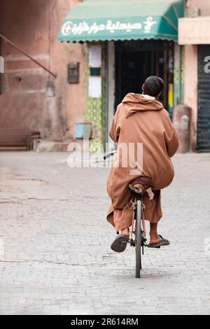 Un homme qui fait du vélo le long d'une allée pavée dans la médina de Marrakech, au Maroc. L'homme porte un Berber Caftan Brown Djellaba avec une cagoule ou qob Banque D'Images