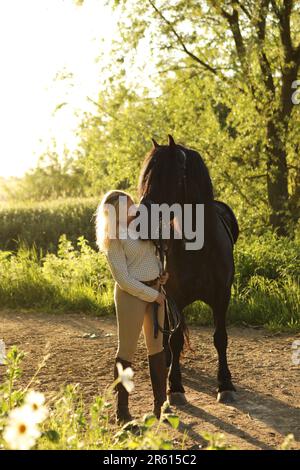 Une femme aux cheveux blonds embrassant un cheval noir une soirée ensoleillée Banque D'Images