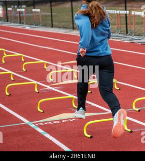 Vue arrière d'une fille de piste d'école secondaire qui se lance sur de petits haies jaunes dans la voie sur une piste rouge pendant l'entraînement de vitesse et d'agilité. Banque D'Images