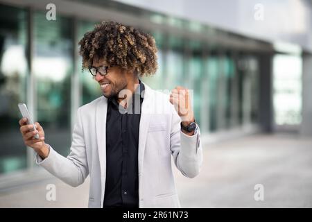 Homme d'affaires noir d'âge moyen et émotionnel tenant un téléphone portable et secouant des poings, en gestant oui, debout à l'extérieur Banque D'Images