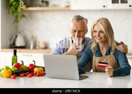 Un couple senior commande des produits d'épicerie en ligne avec un ordinateur portable et une carte de crédit dans la cuisine Banque D'Images