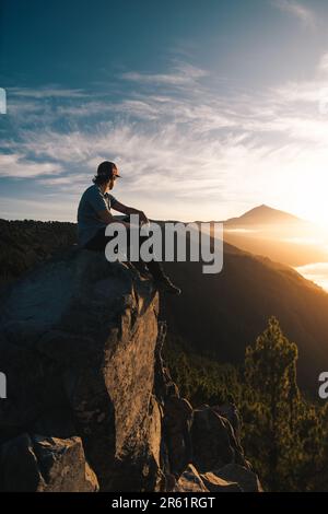 Jeune homme avec cap assis sur un rocher observant le pic de Teide au coucher du soleil pendant son voyage touristique à travers l'île des Canaries de Ténérife. Banque D'Images