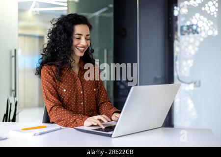 Femme d'affaires hispanique souriante travaillant au bureau avec un ordinateur portable et un casque pour les appels vidéo, femme assise sur le lieu de travail heureuse de travailler avec les clients Banque D'Images