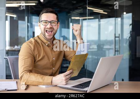 Portrait d'un jeune homme d'affaires heureux assis dans le bureau tenant une enveloppe avec les documents. Un accord financier réussi. Il regarde la caméra, montre un geste de victoire avec sa main Banque D'Images