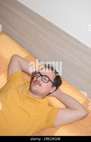 Jeune homme avec des lunettes noires prenant une sieste dans le lit avec les bras sur son cou Banque D'Images