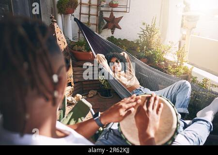 Profitez des battements relaxants. une jeune femme se reposant sur un hamac tout en écoutant son petit ami jouer des tambours. Banque D'Images