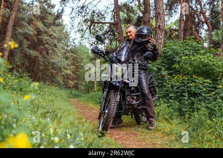 Un cycliste senior qui a pris son casque après avoir fait du motovélo dans la forêt estivale. Homme à la retraite portant des vêtements en cuir assis sur la moto. Passe-temps extérieur Banque D'Images