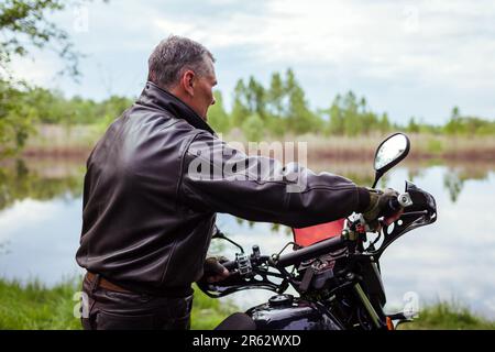 Vue arrière de vélo senior à moto en plein air. Homme tenant la barre de moto par rivière portant des vêtements en cuir. Voyage d'été Banque D'Images