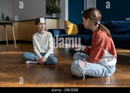 Petit enfant garçon et fille jouant à deviner jeu avec des notes collantes tout en étant assis sur le sol à la maison Banque D'Images