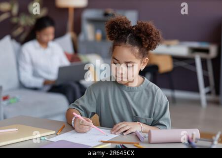 Portrait de fille noire dessinant des images assis à la table à la maison et regardant focalisé avec l'imagination Banque D'Images