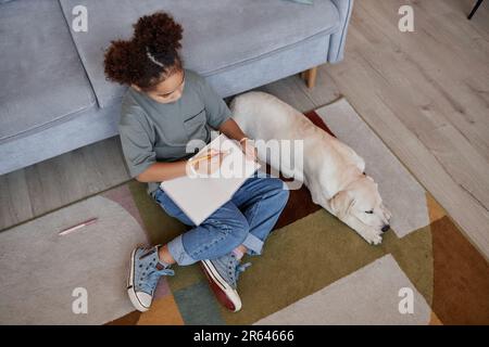 Vue de dessus sur une adolescente noire qui écrit dans un journal tout en étant assise sur le sol à la maison avec un chien blanc, un espace de copie Banque D'Images
