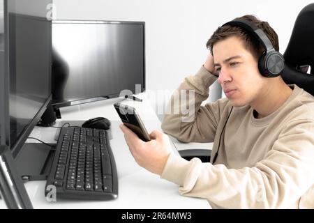 L'étudiant avec un casque tient son smartphone tout en vérifiant les médias sociaux devant l'écran de l'ordinateur au bureau Banque D'Images