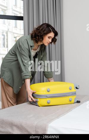 jeune et attrayante femme avec des cheveux de brunette ondulés et tatouage, dans une chemise élégante et pantalon ouvrant valise jaune tout en se tenant près du lit et fenêtre wi Banque D'Images