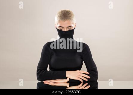 Modèle jeune femme calme avec des cheveux blonds courts couvrant la bouche avec col de turtleneck noir tout en étant assis à une table miroir avec les yeux fermés contre Banque D'Images