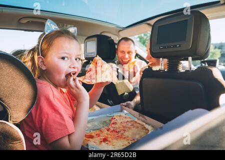 Petite fille à bouche ouverte portrait mangeant de la pizza italienne assis dans une voiture moderne avec mère et père. Des moments de famille heureux, l'enfance, la restauration rapide Banque D'Images